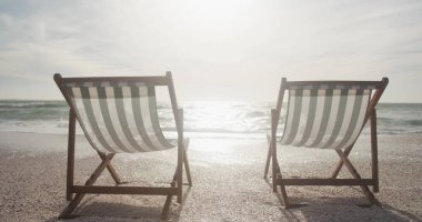İki çizgili iskemle, güneşli bir sahilde, fotokopi alanı olan okyanusa bakıyor. Ayarlar deniz kenarında, tatilde rahatlatıcı bir gün olduğunu gösteriyor..