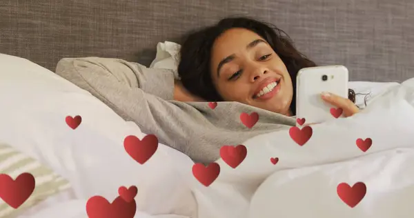 Digital composite of a Hispanic woman lying in bed smiling while texting and digital hearts flying in the foreground