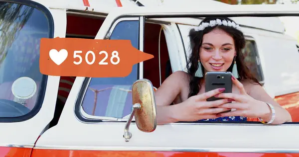 Close up of a Caucasian female hippie leaning on a van door while typing on her phone. Beside her in the foreground is a digital image of a message bubble with a heart icon increasing in count
