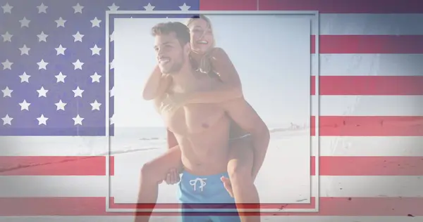 Caucasian woman piggybacking on a Hispanic or Latino man, both smiling. They are enjoying a playful moment on a sunny beach with an American flag backdrop.