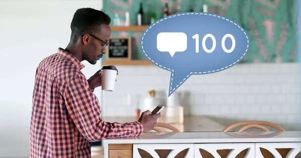 Side view of an African-american man leaning on a kitchen counter sipping on a cup of coffee while texting. Beside him is a digital image of a message bubble with a message icon increasing in count