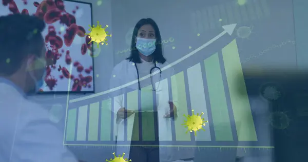Image of yellow virus cells over biracial female doctor in face mask and graphs. Health, medicine, biology, science and covid 19 pandemic concept digitally generated image.