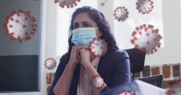 Image of covid 19 cells floating over worried biracial woman wearing face mask in office. Healthcare and protection during coronavirus covid 19 pandemic, digitally generated image.