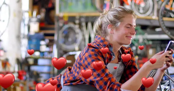 Digital composite of a Caucasian female bicycle mechanic leaning on a table smiling while texting and digital hearts flying in the foreground