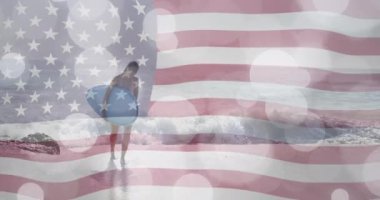 Güneşli sahilde sörf tahtası taşıyan beyaz bir kadının üzerinde Amerikan bayrağı ve ışık lekeleri. Amerika, yaz, tatiller, sağlıklı yaşam tarzı, kutlama ve vatanseverlik, dijital olarak üretilen video.