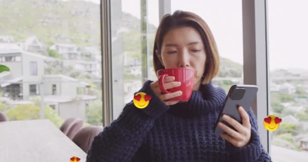 在亚洲女人在家里喝咖啡和使用智能手机时 动画中的情感符号会让人联想到她们 生活方式 通信和家庭生活概念数码视频 — 图库视频影像