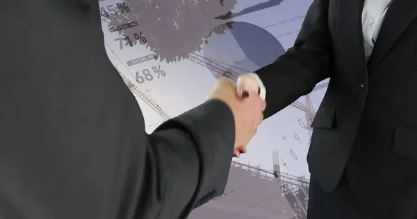 Close up of a handshake between a businessman and a businesswoman. Behind them is a digital image of graphs and statistics running on a background of cranes.
