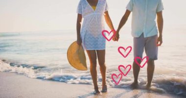 Yaz aylarında kumsalda el ele tutuşan birbirine aşık çiftlerin üzerinde hareket eden kalplerin animasyonu. Aşk, romantizm, sevgililer günü ve tatil konsepti, dijital olarak oluşturulmuş video.