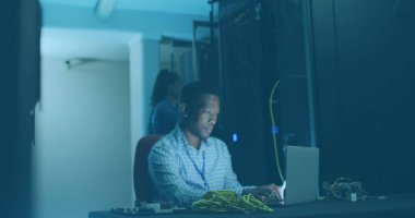 Mavi gömlek giyen, kablo tutan, dizüstü bilgisayarda çalışan Afro-Amerikan bir baba. Gülümsüyor, mavi arkaplanı olan bir sunucu odasında ekrana odaklanıyor