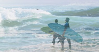 Sörf tahtası tutan, okyanus dalgalarına doğru yürüyen iki adam. İkisi de dalgıç kıyafeti giyiyor, sörf seansına hazırlanıyor.