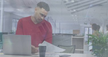 Kısa kıvırcık saçlı, sakallı, kırmızı kazaklı, gazete okuyan melez bir iş arkadaşı. Yanında kahve fincanı olan bir dizüstü bilgisayar kullanırken arka planda başka bir meslektaşım yürüyordu.