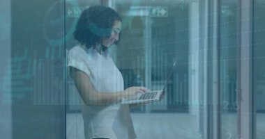 Modern bir ofiste geç saatlere kadar çalışan mutlu bir çift ırklı iş kadını cam kapıya yaslanırken dizüstü bilgisayar kullanıyor.