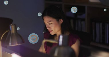 Mor bluz giyen Asyalı genç profesyonel bilgisayar ekranına odaklanıyor. Siyah saçları düzgünce şekillendirilmiş ve loş bir odada kitaplarla birlikte.