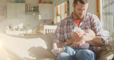 Image of light spots over happy caucasian father feeding baby at home. fathers day and celebration concept digitally generated image. clipart