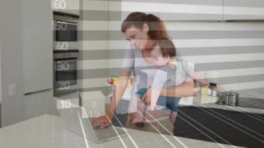 Kucağında çocuk, annesi dizüstü bilgisayar kullanıyor. Mutfakta finansal büyüme çizelgesi var. Ebeveynlik, çoklu görev, uzaktan çalışma, finans, teknoloji, aile
