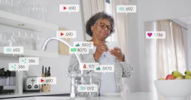 Akıllı telefon kullanan, mutfakta sosyal medya bilgilendirme görüntüsü olan yaşlı bir kadın. Teknoloji, yaşlılar, yaşam tarzı, iletişim, internet, uygulamalar