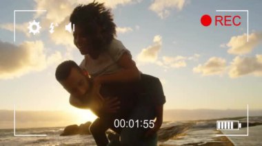 Adam, günbatımı kumsalındaki animasyonu kaydeden bir kadını sırtında taşıyor. Çift, eğlence, yaz, tatil, tasasız, şakacı