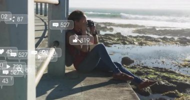 Okyanus kenarında oturup fotoğraf çeken bir kadın hakkında sosyal medya bilgilendirmeleri. Teknoloji, fotoğrafçılık, dijital, tatil, yaşam tarzı, seyahat