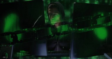 Siyah kapüşonlu giyen, parlayan perdelere odaklanan beyaz bir profesyonel. Loş bir odada birden fazla monitörle çevrili dijital veriler analiz ediliyor