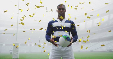 Çift ırklı erkek rugby oyuncusunun üzerine düşen altın konfeti resmi. Kameraya bakıyor, arka planda bir rugby topu tutuyor.