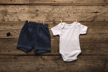 Bir çift bebek kot şortu ve beyaz tulum kırsal ahşap arka planda çamaşır ipine tutturuluyor. Bu büyüleyici gösteri çocukluk masumiyetini ve bebek kıyafetinin basitliğini çağrıştırıyor..