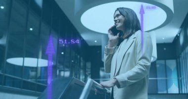 Telefonda konuşan ve gülümseyen, ofis geçmişinde finansal veri görüntüsü olan bir kadın. İş, iletişim, teknoloji, finans, analitik, çalışma