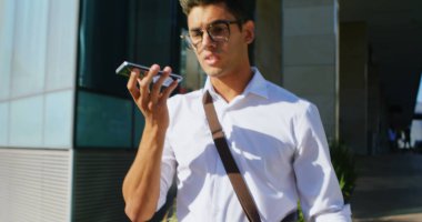 Gözlüklü, akıllı telefondan konuşan melez bir genç. Kısa siyah saçları var, beyaz gömlek giyiyor ve omuz çantası taşıyor.