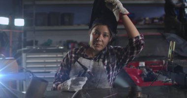Genç bir melez kadın atölyede çalışıyor, elinde kaynak aleti var. Açık kahverengi tenli, koyu renk saçlı ve koruyucu bir önlük giyiyor.