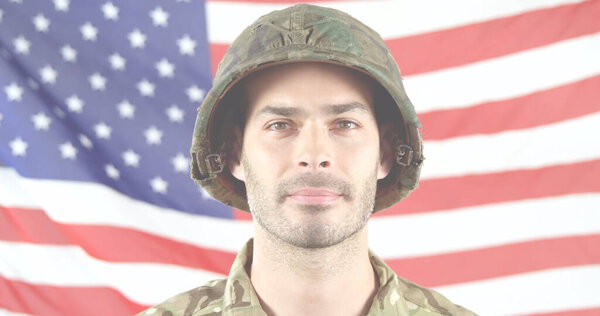 Caucasian male soldier wearing camouflage stands before American flag. He has short dark hair, light stubble, and is wearing helmet