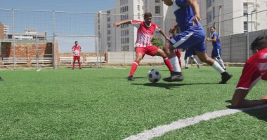 Yeşil sahada futbol oynayan bir grup çift ırklı sporcu. Spor üniformaları ve futbol kramponları giyip topa odaklanıyorlar.