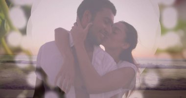Birbirlerine sarılıp öpüşen farklı çiftler. İkisi de beyaz giyiniyor. Koyu renk saçları ve kısa siyah saçları var. İkisi de kahverengi gözlü.