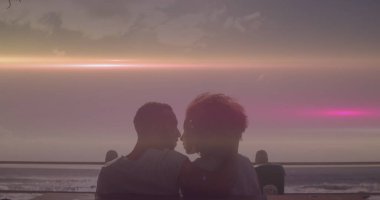 Farklı çiftler deniz kenarında gün batımının keyfini çıkarıyorlar, başları birbirine yakın. İkisinin de saçı kısa, kadın kıvırcık, erkek ise kıyılmış.