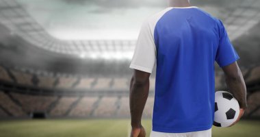 Futbol topu tutan adam stadyum sahasına bakıyor. Mavi forma giyiyor, maça hazırlanıyor.