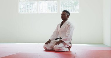 Judo antrenmanından önce beyaz judogi giyen, spor salonunda diz çöküp minderlere eğilen melez bir erkek judoka 'nın yan görüntüsü.