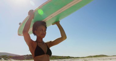 Genç Afro-Amerikan kadın sörfçü düşük açı görünümü gülümseyerek ve plaj 4k de kafasında sörf tahtası taşıyan