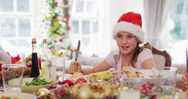 Caucasian girl in santa hat holding fork and knife while sitting on dining table and ready to enjoy lunch together at home. christmas festivity tradition celebration concept clipart