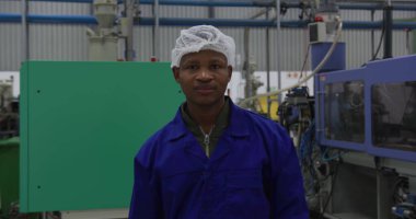 Odaklanmış Afrikalı Amerikalı erkek işçinin portresi yoğun bir fabrika deposunda kameraya bakıyor, saç filesi takıyor ve gülümsüyor.