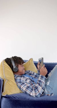 Vertical image of african american boy lying on sofa using smarphone at home. Childhood, communication and domestic life, unaltered. clipart