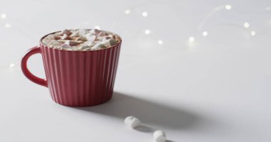 A red mug filled with hot chocolate and marshmallows sits on a table. Festive lights add a cozy ambiance, suggesting a warm holiday treat at home. clipart