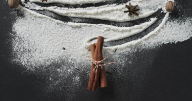 Cinnamon sticks tied with string lie amidst flour on a dark surface. Ingredients are artistically arranged, suggesting baking or festive culinary preparation. clipart