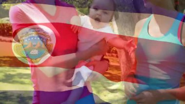 Kosta Rika bayrağı taşıyan bir bebek, parkta duran iki kadın. Aile, ebeveynlik, açık hava, gurur, bağ, aşk