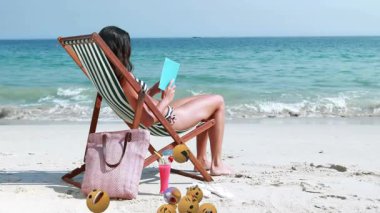 Plaj sandalyesinde dinlenen, yüzen emoji animasyonlu kitap okuyan bir kadın. Tatil, tatil, rahatlama, yaz, dijital, teknoloji
