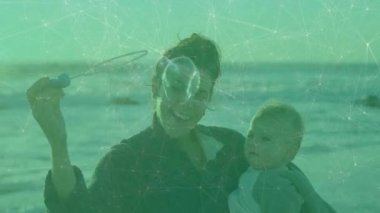 Plajda bebek tutarken şebeke animasyonuyla baloncuk çıkaran bir kadın. Annelik, aile, teknoloji, bağ, neşe