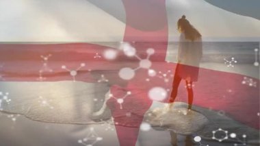 Sahilde yürüyen, elinde moleküler yapı animasyonuyla İngiltere bayrağı tutan bir kadın. Tatil, vatanseverlik, teknoloji, bilim, seyahat, milliyet