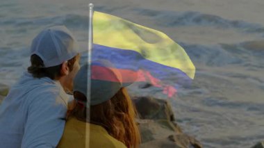 Günbatımı animasyonu sırasında okyanus kenarında oturan insanların üzerinde Kolombiya bayrağı sallıyor. Tropikal, kültür, seyahat, dalga, vatanseverlik, sükunet