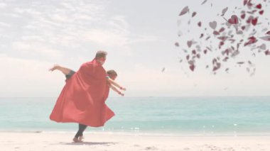 Baba ve çocuk, plajda koşan pelerinli ve kalp animasyonu kaplamalı. Aile, bağlılık, ebeveynlik, süper kahraman, hayal gücü, mutluluk