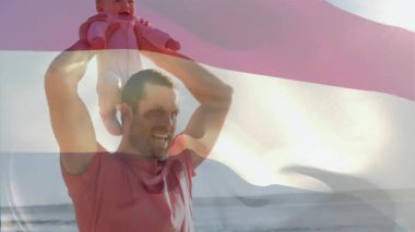 Neşeli bir şekilde çocuğu kaldırıyor, babası arka planda bayrak sallıyor. Vatanseverlik, kutlama, aile, bağ, açık hava, birlik