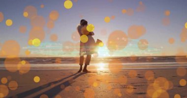 Sahilde kucaklaşan, günbatımı animasyonunun üzerinde parlayan bokeh ışıkları olan çift. Aşk, aşk, deniz kenarı, romantik, kucak, siluetler