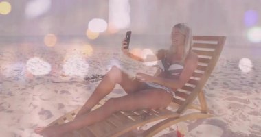 Plaj sandalyesinde selfie çeken, bikinili hafif efektli bir kadın. Tatil, yaz, seyahat, tatil, teknoloji, güneş gözlüğü.