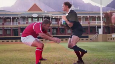 Çimenli alanda top koşturuyor, ragbi oyuncuları büyük tuğla binaların önünde. Spor, rekabet, takım çalışması, aksiyon, atletik, açık hava